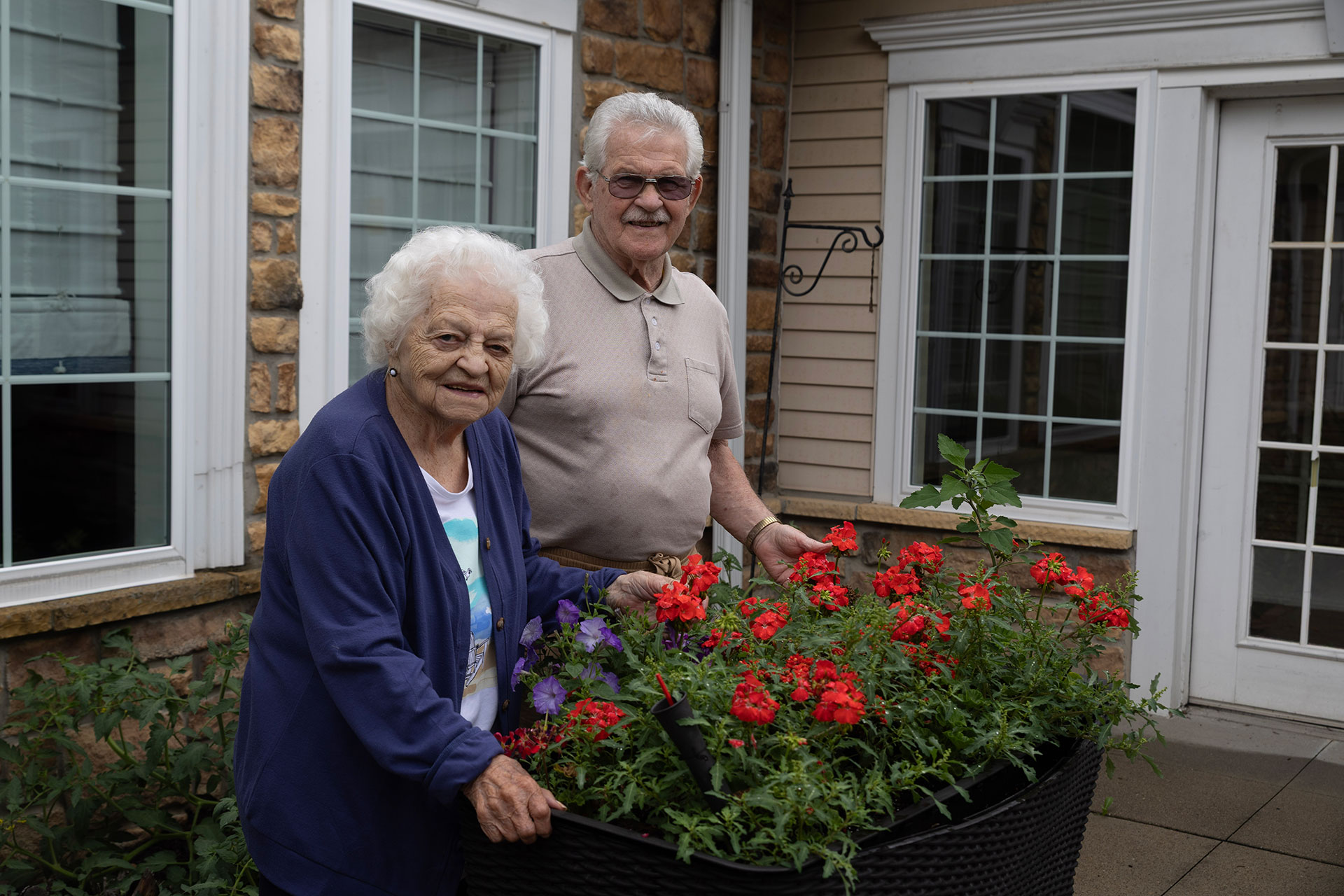 Elderly couple smiling with potted flowers outdoors.