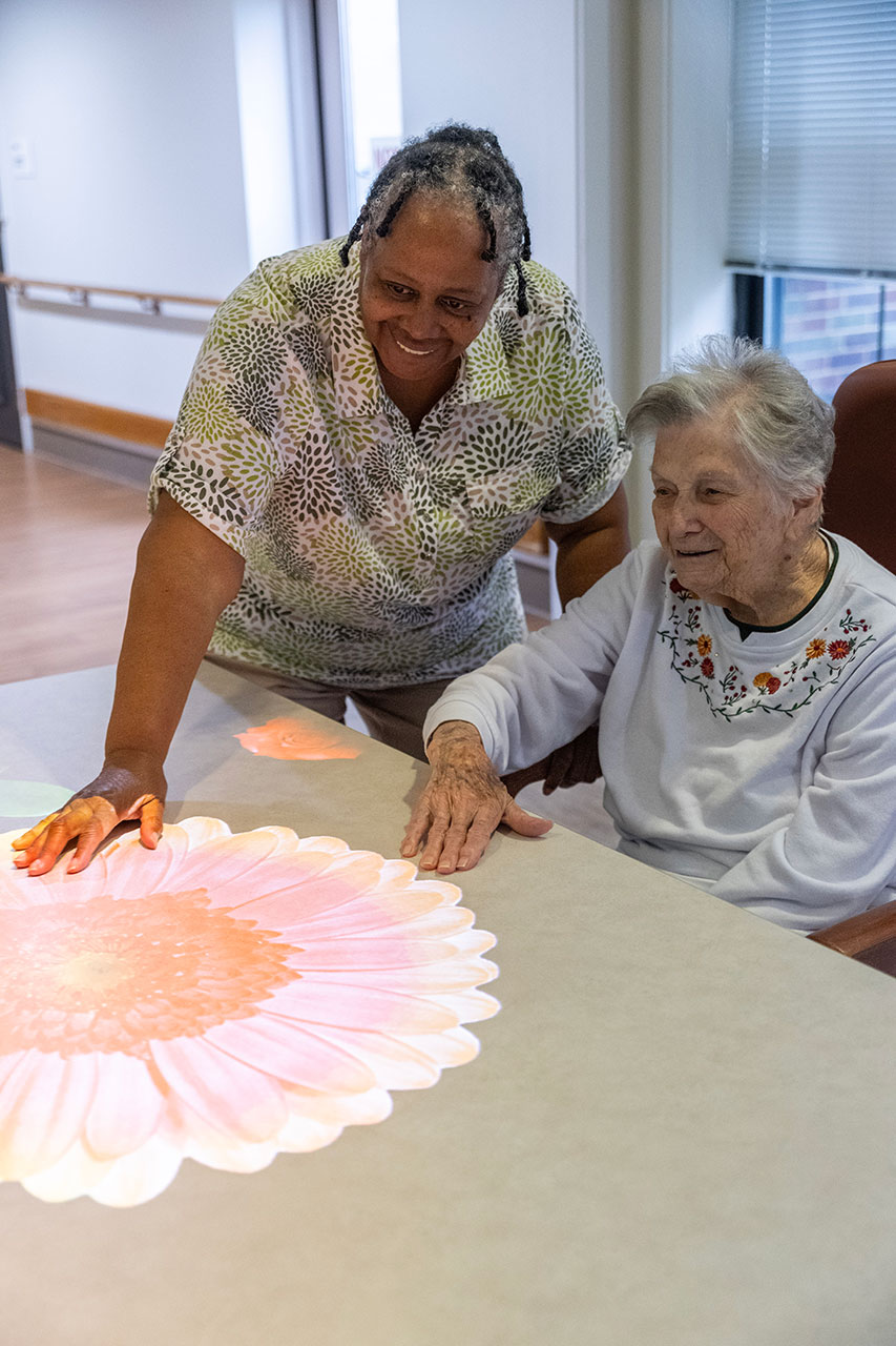 Two women interacting with a projected flower on a table.