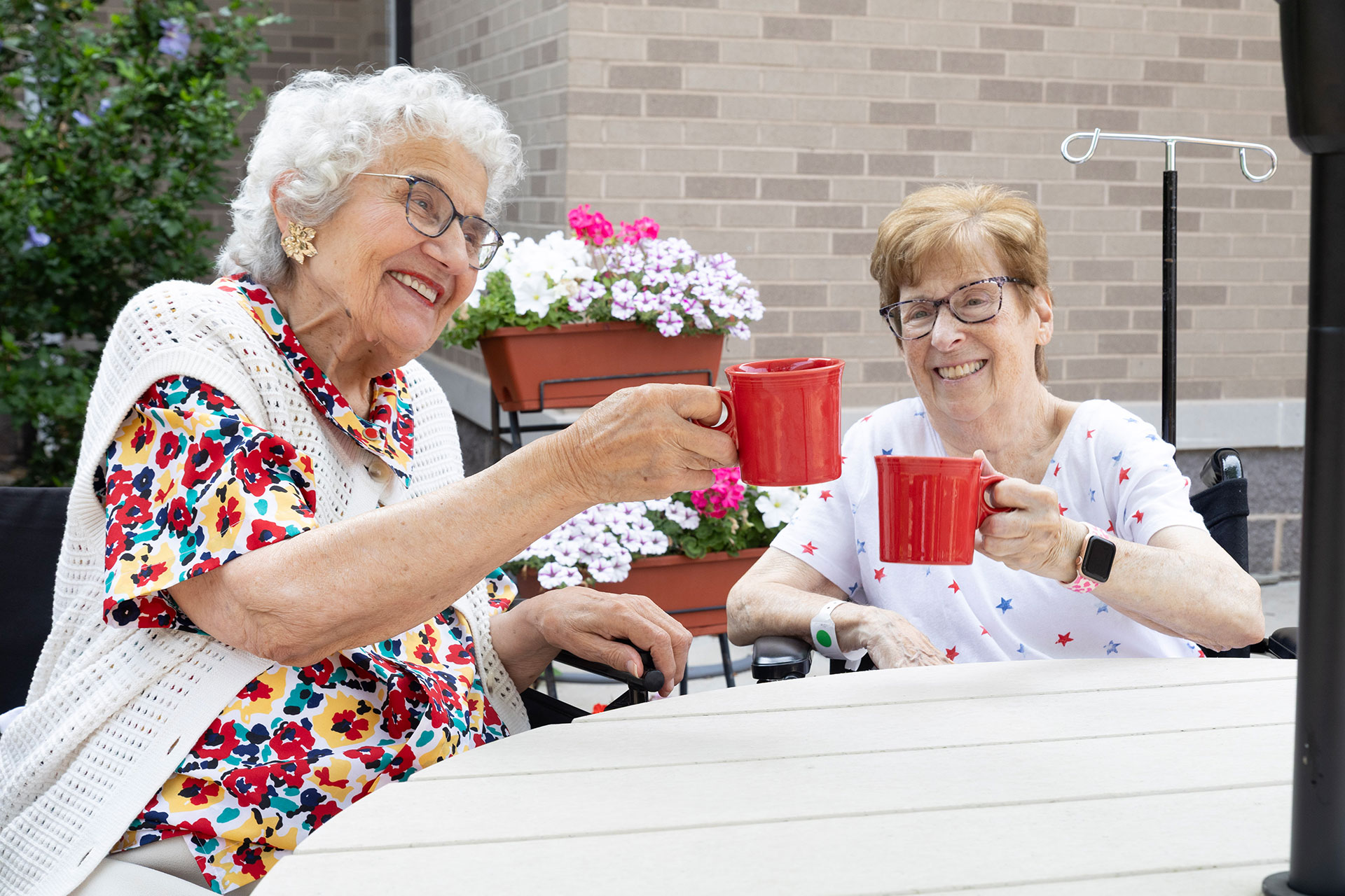 Two elderly women toasting with red mugs outdoors.