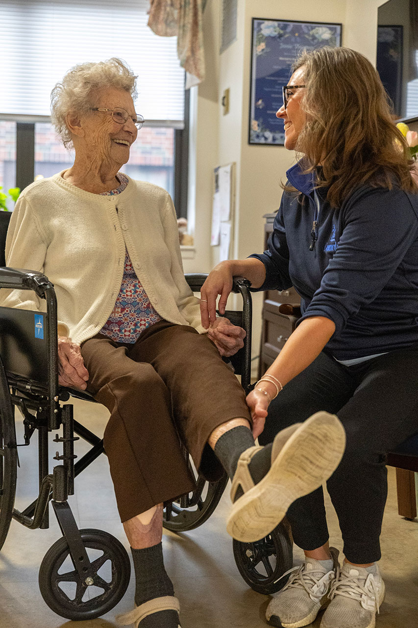A caregiver joyfully interacts with an elderly woman in a wheelchair.