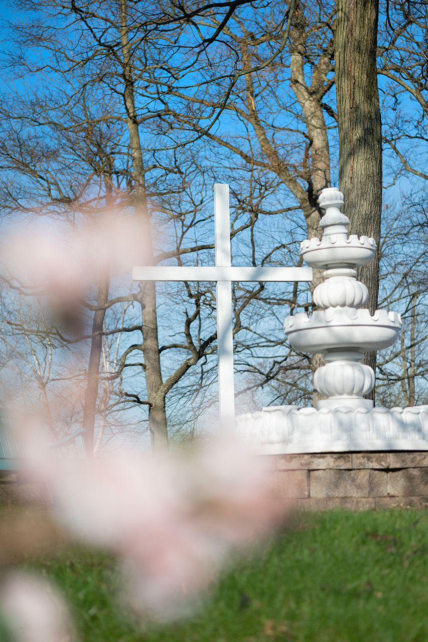 White cross and fountain surrounded by trees and flowers.