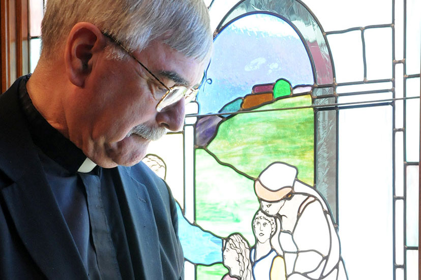 Man in clerical attire beside a stained glass window.