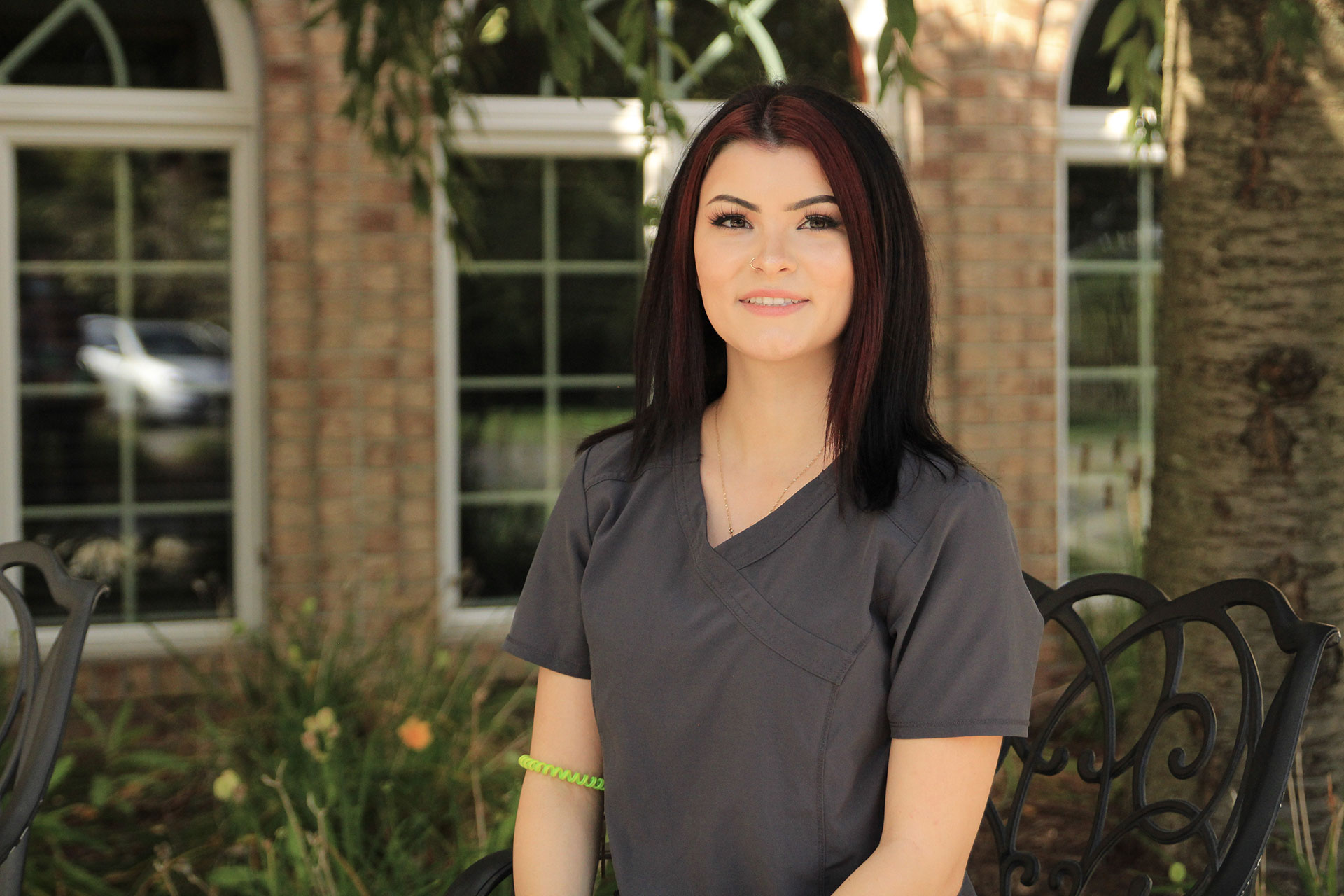 Young woman in scrubs, smiling outdoors near windows.