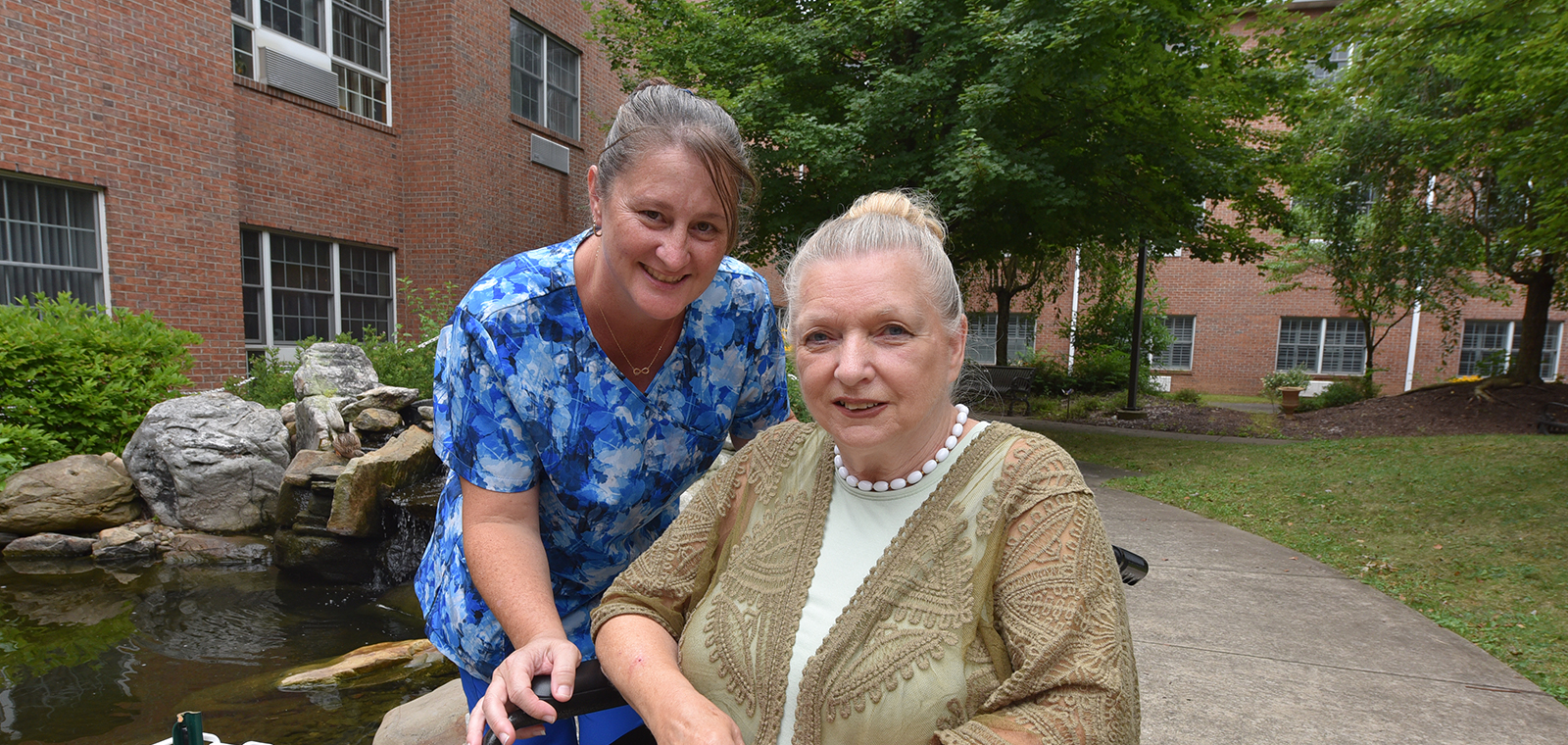 Two women smiling outdoors near a pond and greenery.