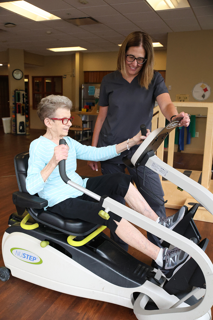 Elderly woman using exercise equipment with assistance.