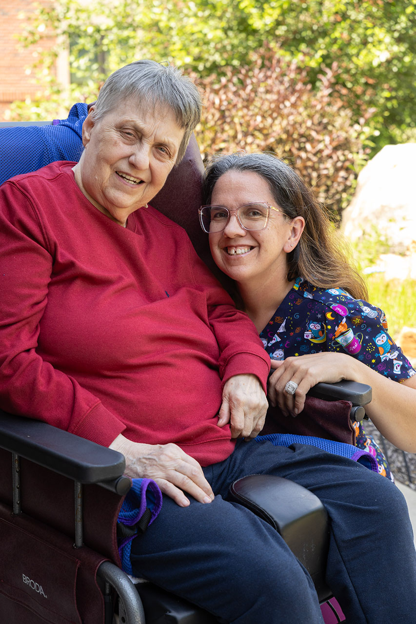 Two women smiling together outdoors, one in a wheelchair.